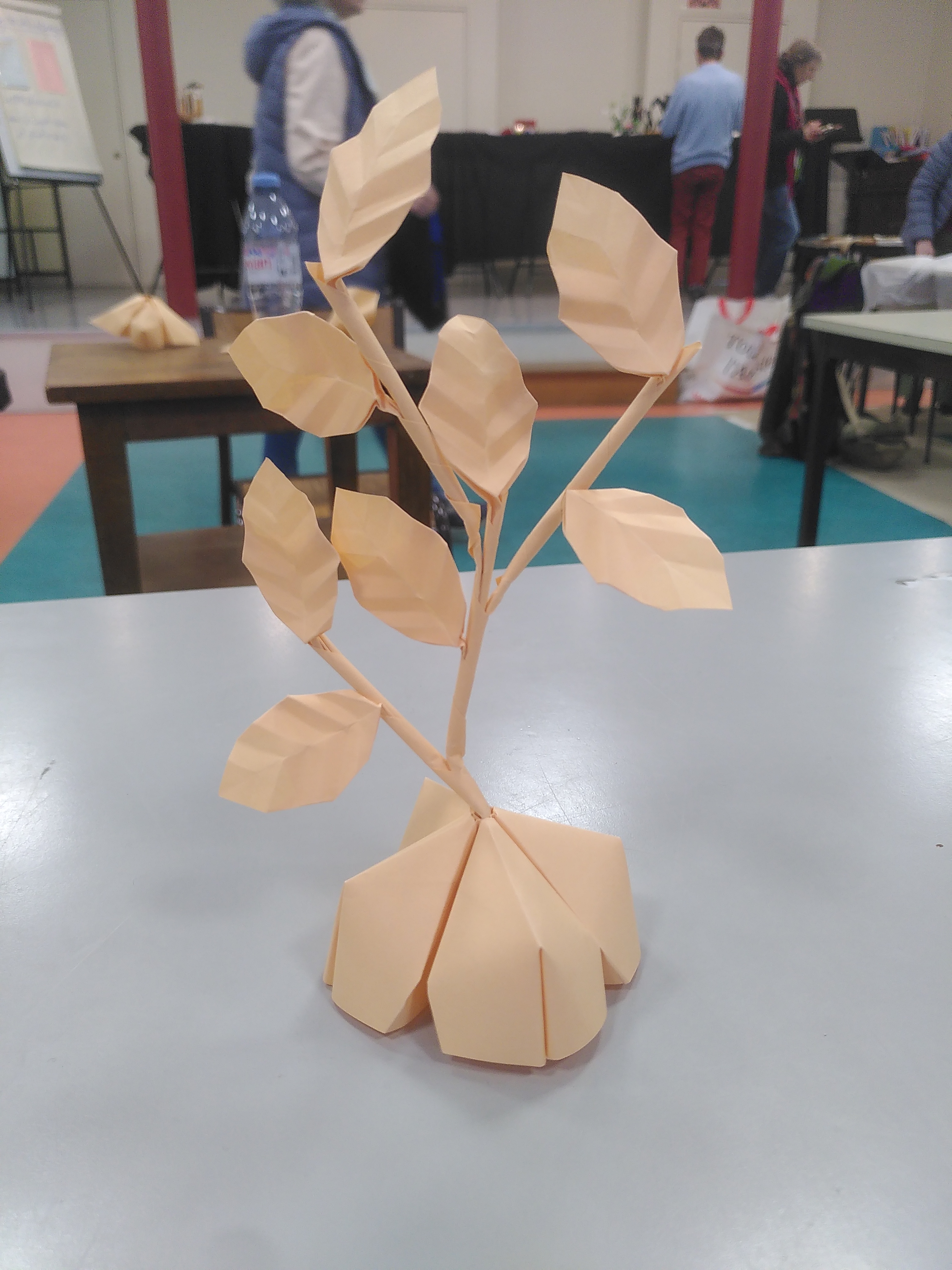 Une composition en origami avec 8 feuilles d'arbre ovales et quatre tiges qui s'emboîtent les unes dans les autres. Le tout est planté dans un socle aussi en origami.