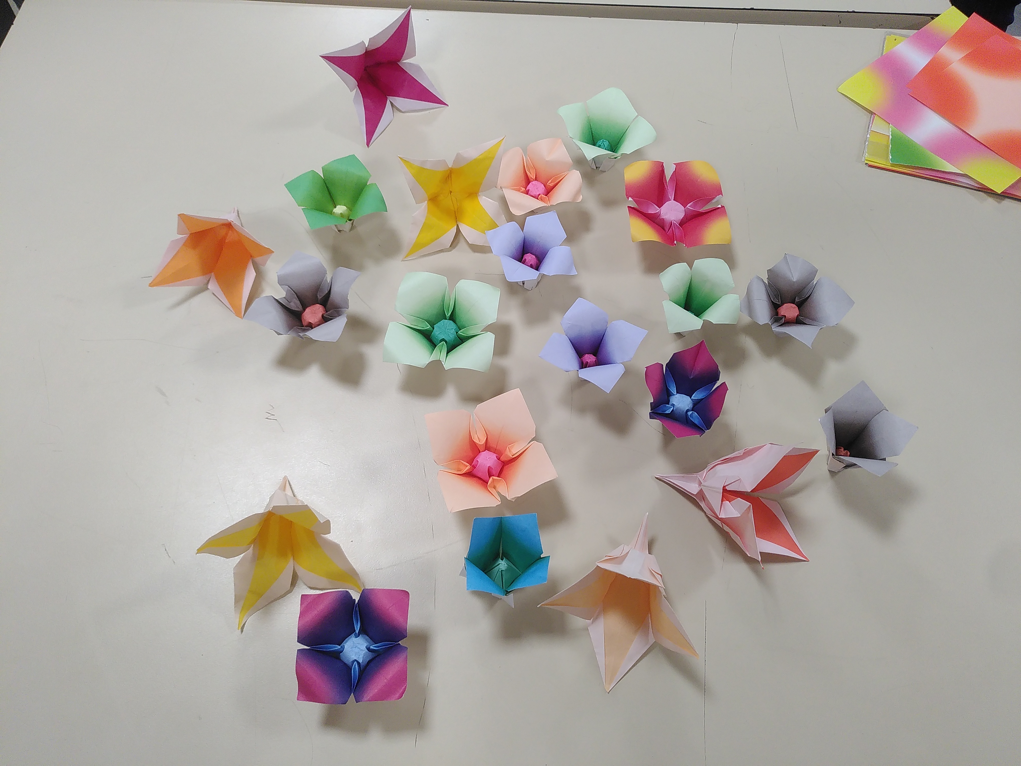 Une vingtaine de fleurs en papier coloré posés sur une table. Toutes ont quatre pétales.