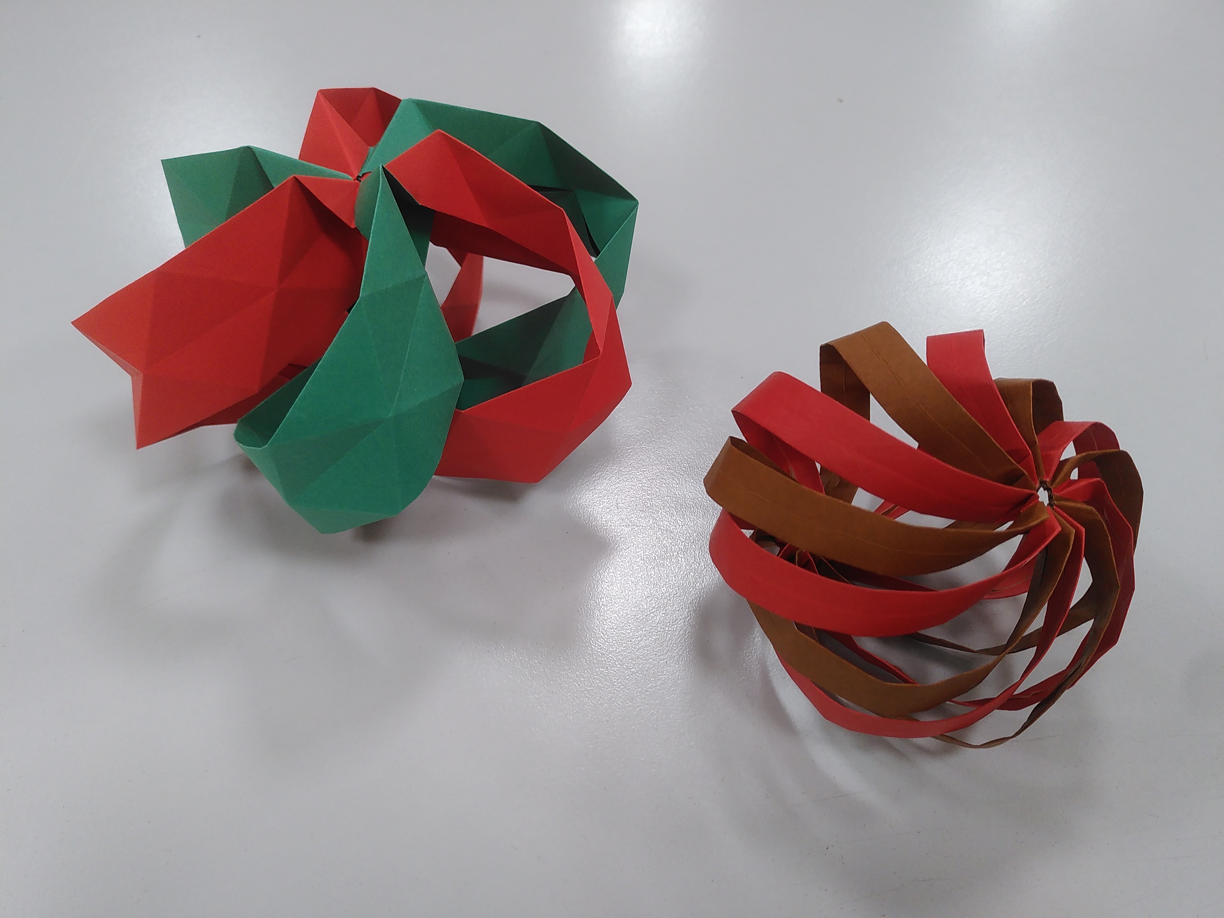 À gauche : le modèle *Seeds in the Wind* en couleurs de Noël (rouge et vert). C'est une sorte de ballon aplati avec trois bandes rouges et trois bandes vertes qui s'enchevêtrent. À droite : un modèle un peu ressemblant mais avec le double de bandes, et deux fois plus fines, et lisses (au lieu d'être quadrillées de plis). C'est un peu plus circulaire, mais un peu aplati quand même, comme un ballon de baudruche rempli d'eau un peu écrasé par son propre poids.