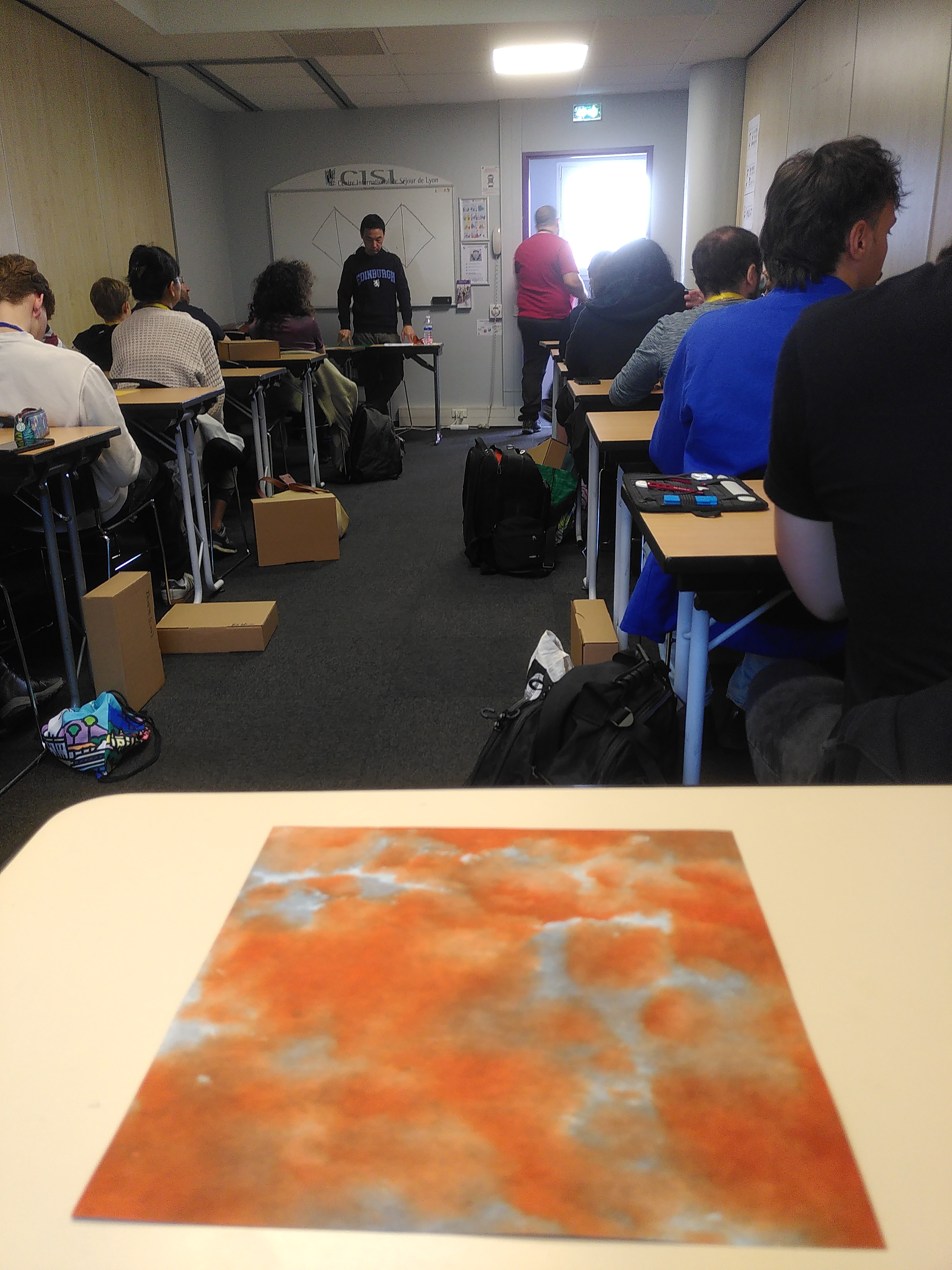 Une salle de classe, je suis assis au fond avec une superbe feuille à motifs orangés et gris sur ma table.