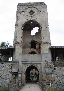 Photo de l’entrée du palais de Krzyżtopór (2008). Un petit pont en bois donne sur une porte arquée ouverte dans une tour. L’arc est encadré à gauche par une croix et à droite par une hache. Chacune mesure près de 3 m. La tour est éventrée, elle a perdu ce qui pouvait être une grande arche à la romaine, haute comme deux arches d’entrées. Au-dessus, un gros œil-de-bœuf. On voit ce qui reste des arches de l’escalier à travers la béance de la tour. Des deux côtés de la tour, muraille avec chemin de ronde couvert par des plaques de zinc pour la conservation. On distingue deux arches du bâtiment central à travers la porte. Le lien renvoie vers la même photo agrandie, sans autre commentaire. 