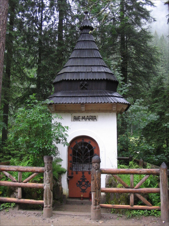 Photo d’une chapelle mariale le long d’un sentier de montagne. Chapelle fermée, blanche porte une inscription «AVE MARYA». Toiture à trois niveaux de tuiles de bois foncé. La porte vitrée a une grille en ferronnerie traditionnelle: une plante à onze branches sort d’un pot marqué des deux marteaux entrecroisées. La chapelle est entourée d’un enclos en bois ouvert par deux sculptures.