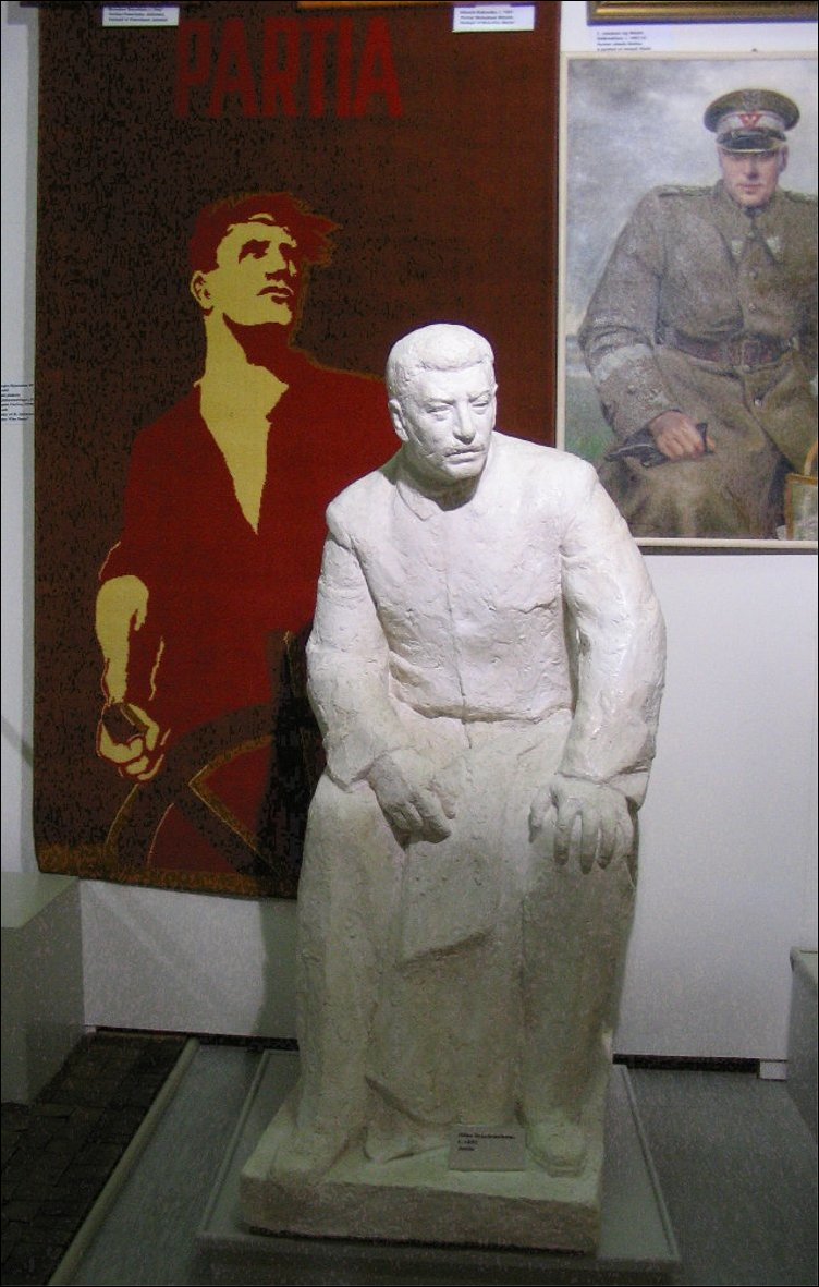 En arrière-plan, une tapisserie représentant le Parti comme un marin barrant un navire et un peinture représentant le maréchal Rokossovski. Au premier plan, statue de Staline.